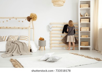 Little girl hanging clothes on handmade rack in scandinavian style bedroom with pillows on carpet