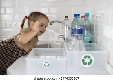 A little girl happily puts plastic bottles into boxes for recycling. In the kitchen, a little girl happily helps sort plastic for recycling. Hive waste concept, environmental friendliness. - Powered by Shutterstock