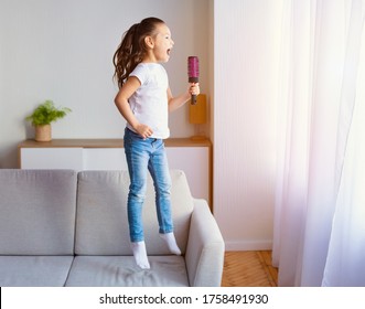 Little Girl Having Fun At Home Jumping On Couch And Singing Holding Hairbrush Like Microphone Indoors. Child's Leisure Concept.