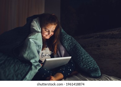 Little Girl Having Fun Watching, Playing And Listening To Stories On Tablet Computer. Child Having Fun Before Bedtime Laying Under Duvet Using Tablet