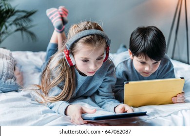 little girl in headphones and boy using digital tablets while lying on bed