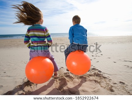 Little girl and her brother sitting on inflatable hoppers and bouncing on the beach on a bright, sunny day
