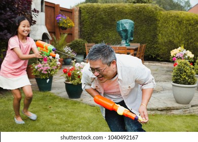 Little Girl And Her Father Are Having A Water Fight In The Garden With Water Pistols. The Girl Is Getting The Dad And He Is Crouching And Trying To Run Away.