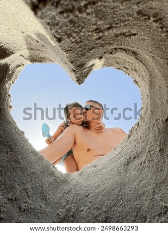 Little girl with her father on the beach photographed from a hole in the sand made in the shape of a heart. Summer family photo from a happy holiday.
