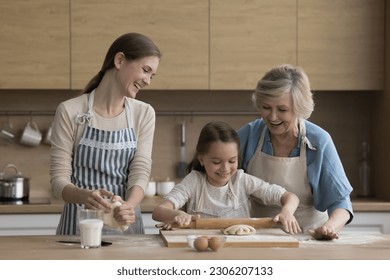 Little girl her grandmother and loving young mom wear aprons cooking together in kitchen, flattening prepared dough making homemade delicious buns, enjoy communication, pastime together and cookery - Powered by Shutterstock