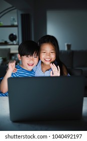 Little Girl And Her Younger Brother Are Using The Laptop To Video Call Their Family. They Are Happily Waving To The Laptop.