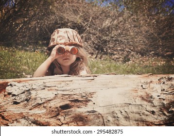 A little girl is hiding behind an old log in the woods with a camouflage hat and binoculars searching and playing for an imagination or exploration concept.