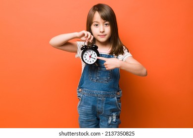 Little Girl Holding An Alarm Clock And Pointing At The Current Time While Looking Serious In A Studio