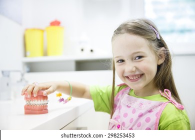 Little Girl Holding An Artificial Model Of Human Jaw With Dental Braces In Orthodontic Office, Smiling. Pediatric Dentistry, Aesthetic Dentistry, Early Education And Prevention Concept.  