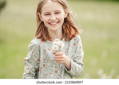 Little girl holding blowballs flower in hands in blossom field and smiling. Cute child kid with dandelions at nature outdoors portrait