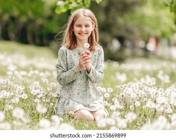 Little girl holding blowballs flower in hands in blossom field and smiling. Cute child kid with dandelions at nature
