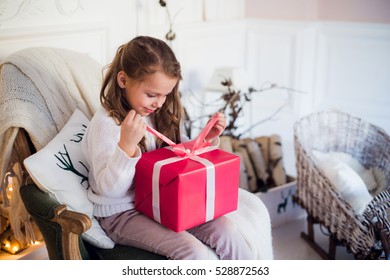 Little Girl Holding A Christmas Gift Box. Cute Kid Sitting On The Chair And Unpacks Xmas Presents Over The Fireplace At Background