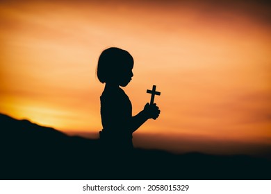 niñita sosteniendo la cruz en las manos y rezando con la luz del fondo de la puesta de sol, concepto de silueta cristiana.