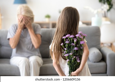 Little girl holding flowers making surprise for grandmother, congratulating her with birthday, excited granny sit with eyes closed, small grandchild prepare gift standing with bouquet in hands