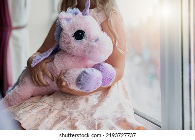 Little girl holding her soft toy unicorn while sit near window