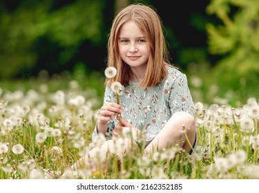 Little girl holding white blowballs flower in hands and looking at camera in blossom field. Cute child kid with dandelions at nature