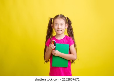 a little girl holds a book and pencils isolated on a yellow background
