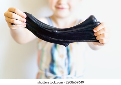 A Little Girl Holds In Her Hands And Stretches The Black Slime. A Child Is Playing With A Slippery, Sticky Toy. A Child Stretching Mucus. Children's Hands With Shiny Black Slime Close-up. Fun Games