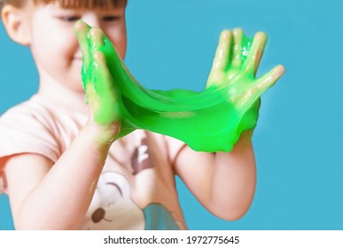 A Little Girl Holds And Stretches In Her Hands A Green Slime On A Blue Background. A Child Is Playing With A Slippery, Sticky Toy. Chewing Gum In Children's Hands. Entertainment For Children.