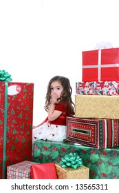 Little Girl In Holiday Dress Sitting In A Huge Pile Of Christmas Presents With Her Hand Over Her Mouth.