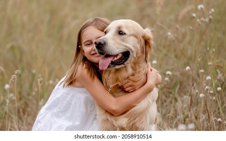 Little girl hugging golden retriever dog in the field in summer day together. Cute child with doggy pet portrait at nature
