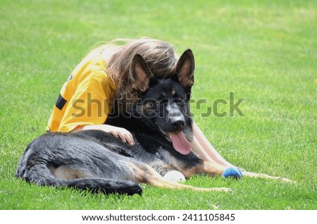 Little girl hugging her big German Shepher dog, sitting on the grass outside in the backyard on a sunny summer day. Dog looking and alert, girl with head buried on dogs head and hugging er dog.