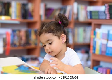 Little Girl Indoors In Front Of Books. Cute Young Toddler Sitting On A Chair Near Table And Reading Book. Child Reads In A Bookstore, Surrounded By Colorful Books. Library, Shop, Shelving In Home.