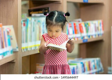 Little Girl Indoors In Front Of Books. Cute Young Toddler Standing Reading Book. Child Reads In A Bookstore, Surrounded By Colorful Books. Library, Shop, Shelving In Home