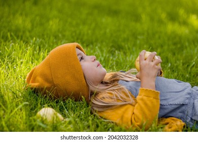 A Little Girl In A Jacket And Hat Lies On The Lawn And Eats Pears In The Fall.