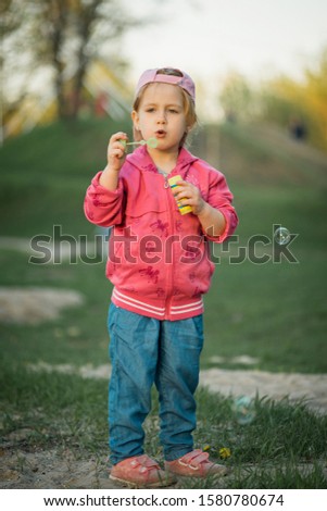 A little girl in jeans and a pink jacket plays on the river bank in the sun and blows soap bubbles