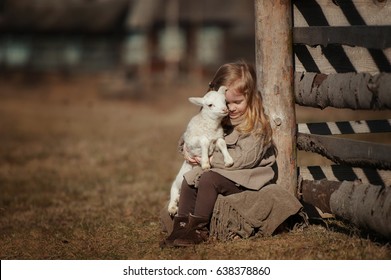 little girl with lamb on the farm. She sits by the fence and hugs the lamb.
