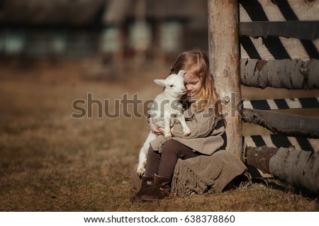 little girl with lamb on the farm. She sits by the fence and hugs the lamb.