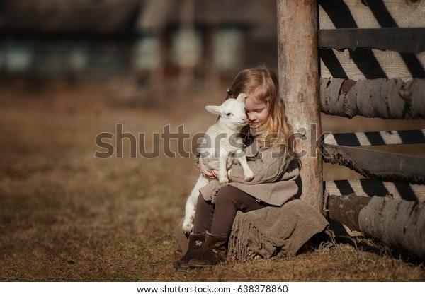 little girl with lamb on the farm. She sits by the\
fence and hugs the lamb.