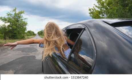 Little Girl Leaning Out Of Car Window And Holding Her Hand Out While Riding Through Country Road. Small Child Putting Her Arm Out Of Open Window Moving Auto To Feel The Breeze. Rear View Slow Motion