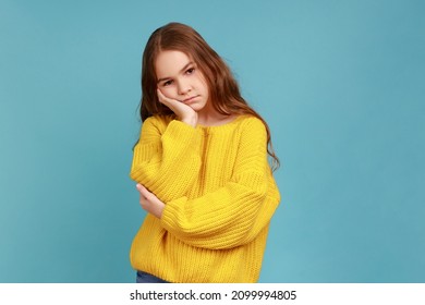 Little girl leans on hand, looking depressed, child with indifferent gloomy expression has no energy, wearing yellow casual style sweater. Indoor studio shot isolated on blue background.