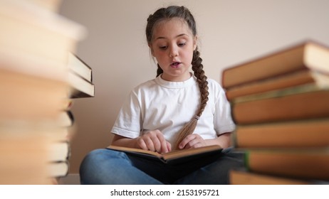 Little Girl In The Library Reading A Book. Education School Kid Science Concept. A Child Sits On The Floor Stacks Lifestyle Of Books Flips Through The Pages Reads Studies. Daughter Reading A Book