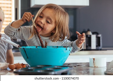 Little girl licking spoon while mixing batter for baking in kitchen  and her brother standing by. Cute little children making batter for baking.