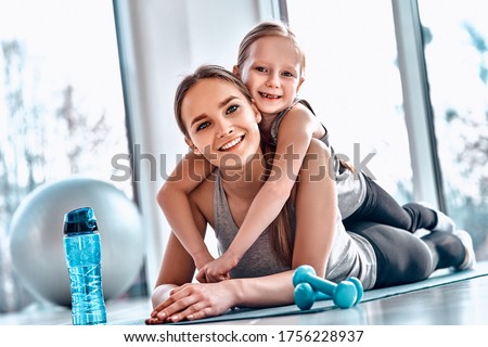 A little girl lies on her mother's back while resting after workout in gym atmosphere with dumbbells, mat, fitness ball, bottle of water on the floor. Healthy family leisure concept