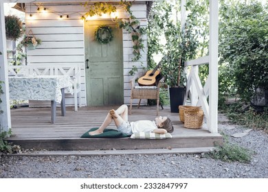 Little girl lies on pillows on terrace of rustic house. Dreamy cute kid put her hands under head and looks up at sky. Retro interior with wooden table, wicker chair, baskets and light bulb garland. - Shutterstock ID 2332847997