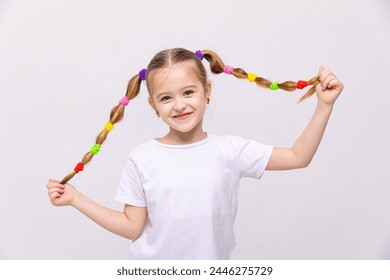 The little girl lifts her braids of hair up, with multi-colored elastic bands, and smiles broadly. Hairstyles for little girls. White isolated background. - Powered by Shutterstock