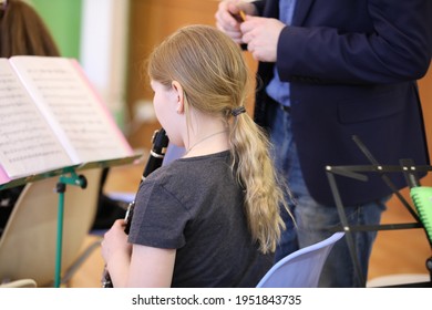 A Little Girl With Long Blonde Hair Pulled Back In A Ponytail In The Classroom In Music Class Looks At The Notes And Plays The Clarinet To The Teacher Standing Next To Her. School Education Concept