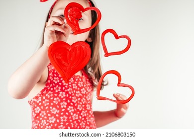 Little girl with long curly hair in red clothes draw hearts with paint on transparent glass smiling happily on white background. Family, love, holidays, birthday, Mother's day concept. Copy space.