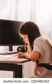 Little Girl With Long Hairs Sitting At Table, Holding Pen, Writing, Learning At Home. Teenager Studying Indoors At Room With Modern Black Chair. Home Education