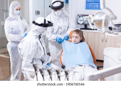 Little Girl Looking At Dentist While Assistant Is Putting Her A Bib Before Dental Examination Uring Coronavirus Pandemic. Stomatologist During Covid19 Wearing Ppe Suit Doing Teeth Procedure Of Child