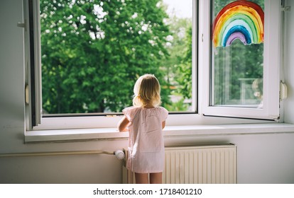 Little Girl Looks Out Open Window At Spring Time. Painting Rainbow On Window. Open Window In Room With Green Trees On Background. Kids Leisure At Home. Support During Quarantine. Feeling Fresh Air.