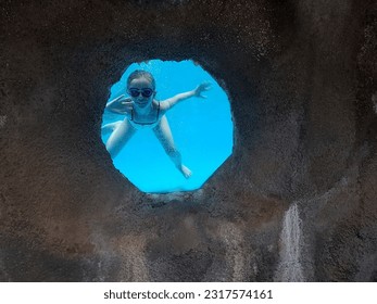A little girl looks through a window under the water of a swimming pool