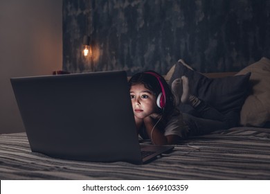 Little Girl Lying In Bed Focused While Watching A Movie On A Laptop In The Darkness, Has A Pink Headset, Child And Technology Concept, Copy Space For Text