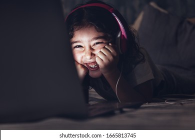 Little Girl Lying In Bed Laughing While Watching A Movie On A Laptop In The Darkness, Has A Pink Headset, Child And Technology Concept, Copy Space For Text