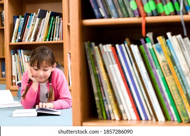 Little girl lying down on the floor in the library and reading an open book in front of her with a blurred bookshelf in the foreground - Powered by Shutterstock