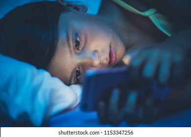 Little Girl Lying On Bed And Using Smart Phone Late At Night. Copy Space. Close Up.